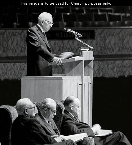 President Joseph Fielding Smith standing at the pulpit and speaking at the Manchester area conference in 1971, with three General Authorities sitting nearby.