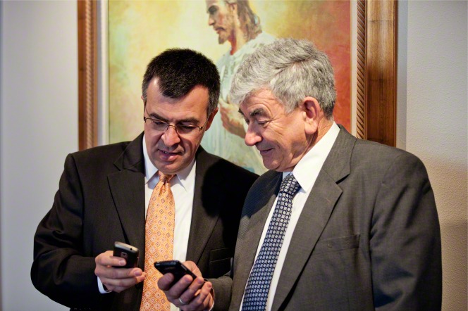 Two older men stand in a meetinghouse next to a picture of Christ and look at the smartphones in their hands.