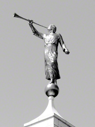 A black-and-white close-up photograph of an angel Moroni statue standing atop a temple spire on a clear day.