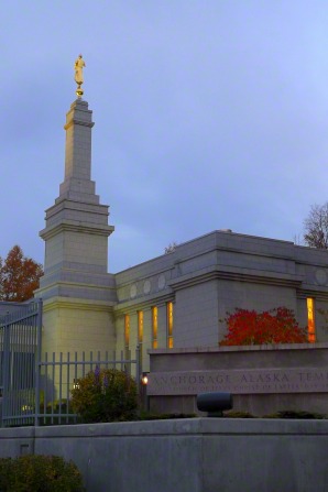 A view of the granite sign on the grounds of the Anchorage Alaska Temple, with the spire and angel Moroni in the background on a fall evening.