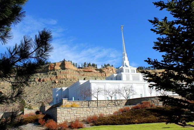 The Billings Montana Temple, with a large rock wall in the foreground and tall cliffs in the background.