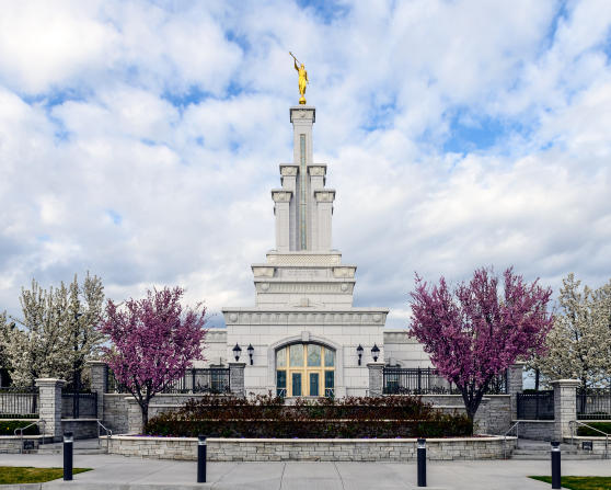 The front of the Columbia River Washington Temple on a spring day, with pink blossoms on the trees and a blue sky full of clouds behind the spire.
