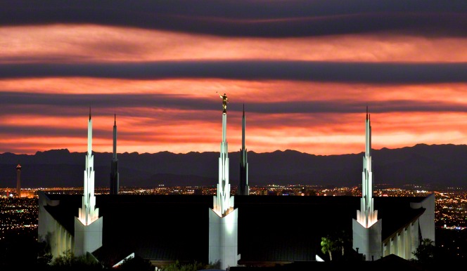 A view of the Las Vegas Nevada Temple illuminated in the evening, with an orange sky in the background and the city lights in the distance.